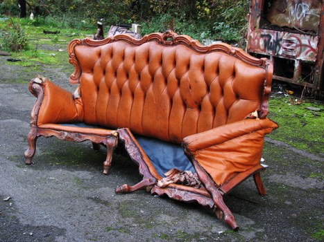 A damaged antique-style sofa with a wooden frame and orange-brown leather upholstery, located outdoors on a paved surface. The sofa's backrest is tufted with button detailing, and the wooden frame features ornate carved detailing. The right armrest is notably torn, exposing foam and internal padding, and the leather surface shows signs of wear and creasing. The seat cushion is displaced and partially hanging off, revealing internal stuffing. The furniture appears to be abandoned in an outdoor environment with greenery and trees in the background, alongside a graffiti-covered storage unit or container on the right. The surrounding area is mossy and weathered, indicating it has been left outside for some time. This image aligns with rubbish removal services offered by Commercial Waste Removal Elephant and Castle, as it depicts a piece of discarded furniture needing clearance.