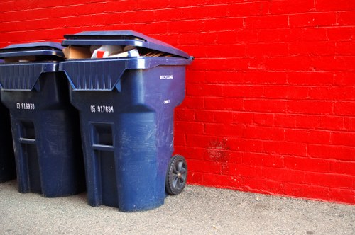 Two large blue wheelie bins made of durable plastic with textured surfaces and black lids are positioned on a gravel pavement next to a red brick wall. One bin appears to have some cardboard visible inside, indicating rubbish collection. The bins are placed side by side, with the one on the right slightly overlapping the wall, which shows a uniform, painted red finish. The environment suggests an exterior setting, possibly a residential or commercial building in Elephant and Castle. The bins' wheels are visible, and they are placed close to the building's exterior, ready for rubbish removal. The overall scene is well-lit, showing clear details of the textured surfaces and contrasting colours between the blue bins and red brick wall, aligning with waste management services like those provided by Commercial Waste Removal Elephant and Castle in central London areas focusing on rubbish collection and disposal.