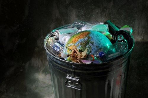 A close-up view of a cylindrical metal rubbish bin with a shiny, reflective surface and a secure latch, filled with a mixture of waste objects. Inside the bin, there is a small, detailed globe showing landmasses and oceans, alongside crumpled brown paper, clear plastic bottles, aluminum cans, and other miscellaneous plastic containers. The background features a dark, textured surface that highlights the contents of the bin, emphasizing the variety of materials discarded, including plastics and paper. The scene suggests an environmentally conscious approach to waste management, aligning with rubbish removal services in the Elephant and Castle area. The lighting appears subdued, with some reflections on the metallic surface of the bin, drawing focus to the diverse waste materials stored within.