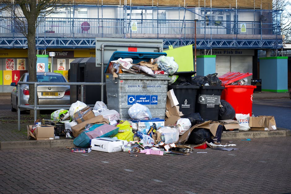An outdoor scene featuring a large pile of mixed waste and rubbish bags accumulated beside a recycling and waste container on a paved area. The waste includes various black bin bags, cardboard boxes,